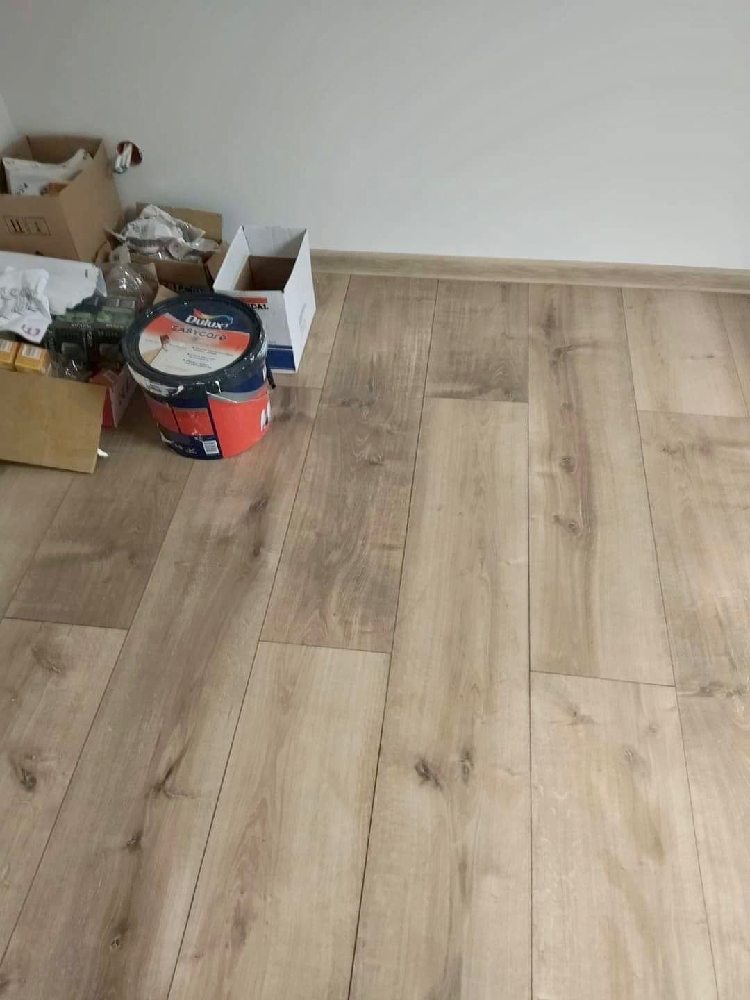 Świeżo położona podłoga z paneli imitujących drewno w jasnym odcieniu, z widoczną puszką farby Dulux Easycare i kartonami w rogu pokoju, wskazującymi na trwający remont.