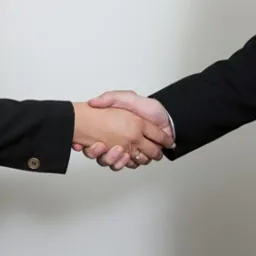 Uścisk dłoni dw&oacute;ch os&oacute;b w ciemnych garniturach na tle jasnej ściany, widoczny pierścionek na palcu jednej z os&oacute;b.