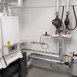 RAD-SERWIS - Dwa białe kotły gazowe z otwartymi panelami, widoczne rury, zawory i manometry, całość zaizolowana termicznie folią aluminiową, żółto-zielony pasek na ścianie.