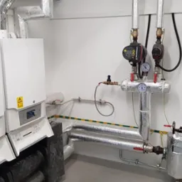 Dwa białe kotły gazowe z otwartymi panelami, widoczne rury, zawory i manometry, całość zaizolowana termicznie folią aluminiową, żółto-zielony pasek na ścianie.