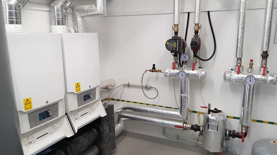 Dwa białe kotły gazowe z otwartymi panelami, widoczne rury, zawory i manometry, całość zaizolowana termicznie folią aluminiową, żółto-zielony pasek na ścianie.