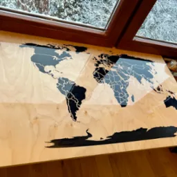 Drewniany blat z wkomponowaną mapą świata z żywicy epoksydowej, widok z góry, obok okno z widokiem na ośnieżone drzewa.