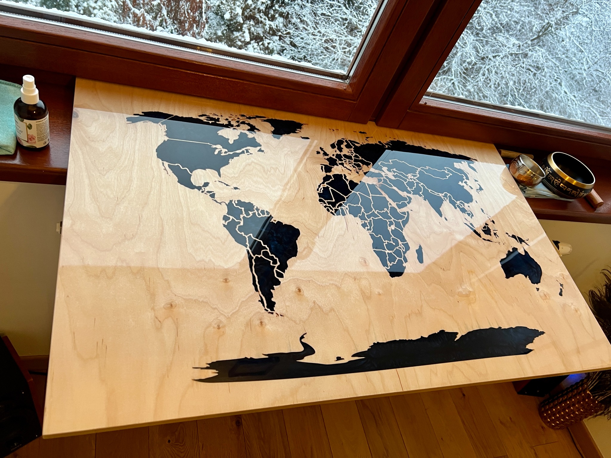 Drewniany blat z wkomponowaną mapą świata z żywicy epoksydowej, widok z góry, obok okno z widokiem na ośnieżone drzewa.