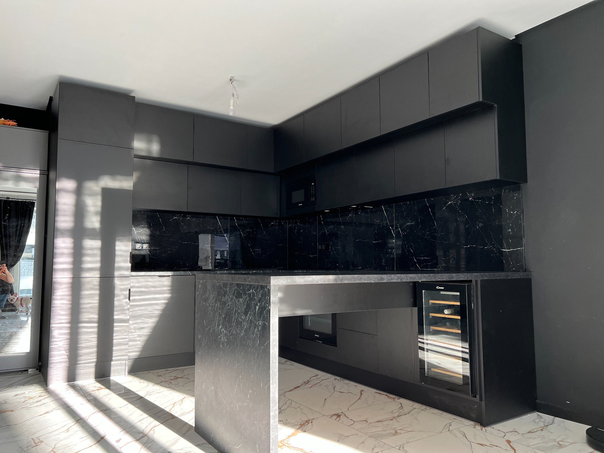 Nowoczesna, czarna kuchnia z wyspą i marmurowym blatem w mieszkaniu we Wrocławiu, z widoczną chłodziarką do wina i oświetleniem podszafkowym.