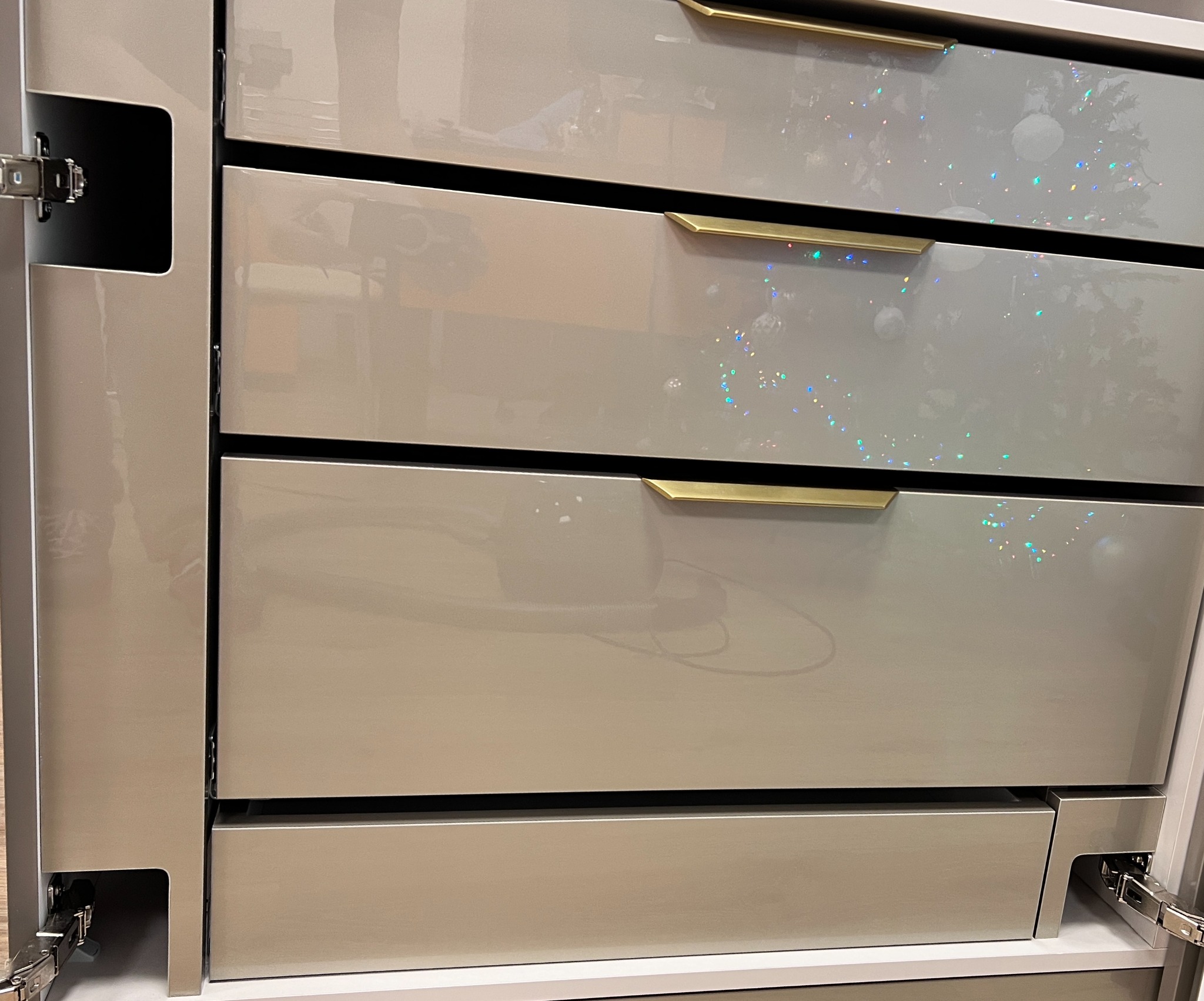 Szare, lakierowane fronty szuflad z poziomymi, złotymi uchwytami, widoczne zawiasy i metalowe elementy konstrukcyjne.