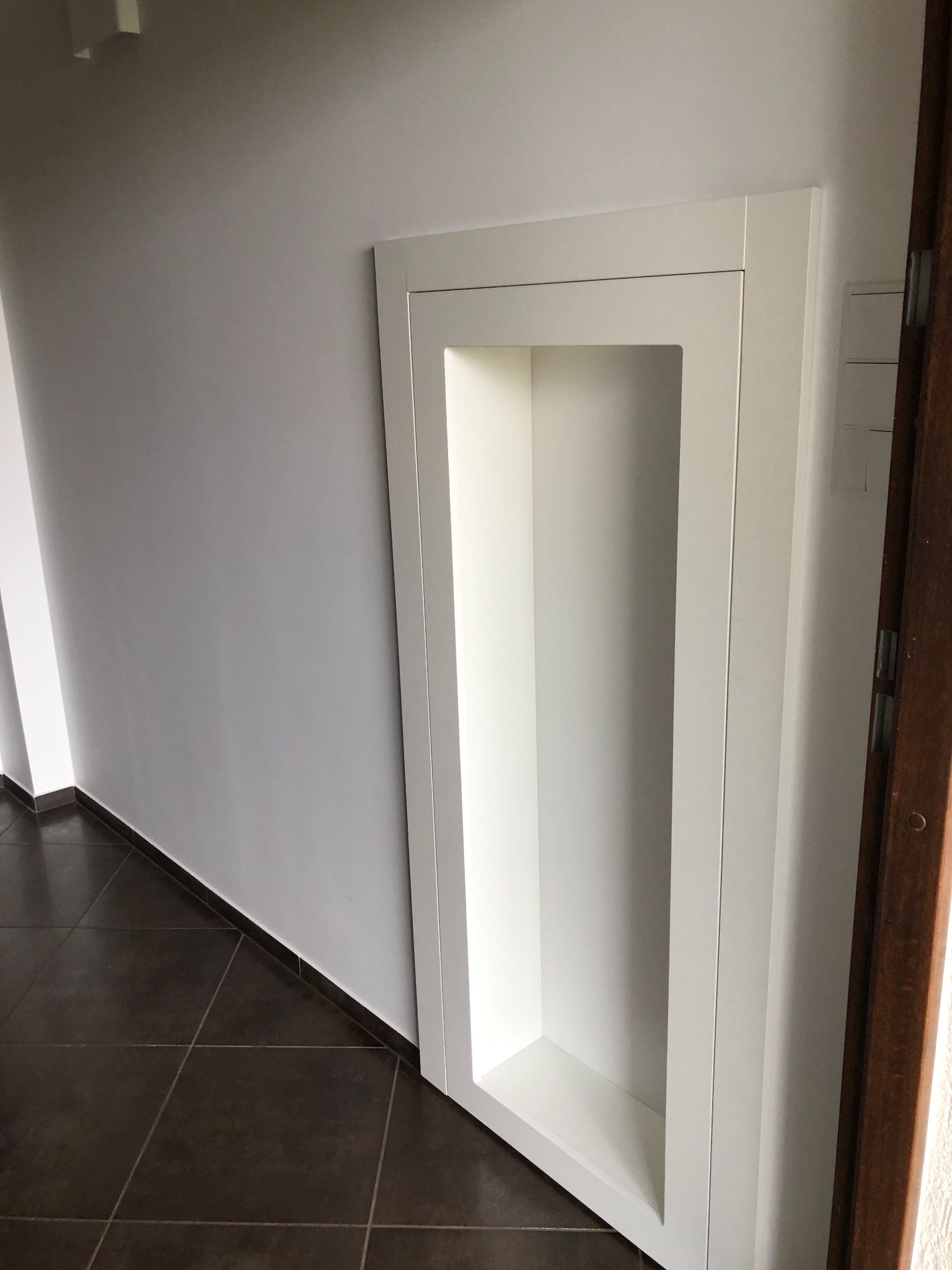 Narożna wnęka ścienna z białym obramowaniem, widok na brązową podłogę z płytek i fragment drewnianych drzwi po prawej stronie.