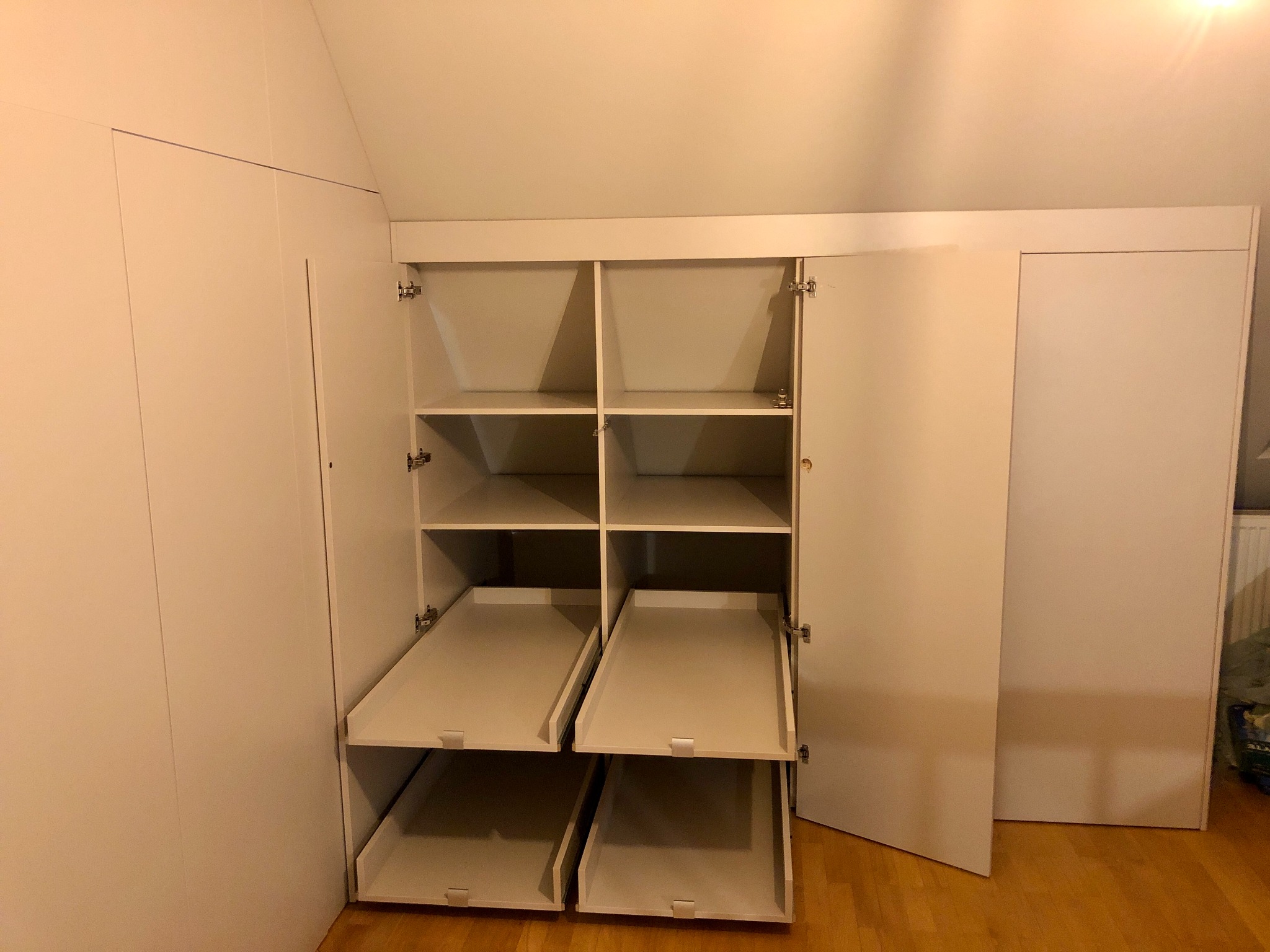 Biała szafa wnękowa z otwartymi drzwiami i wysuwanymi półkami, widoczna na tle drewnianej podłogi i białych ścian.