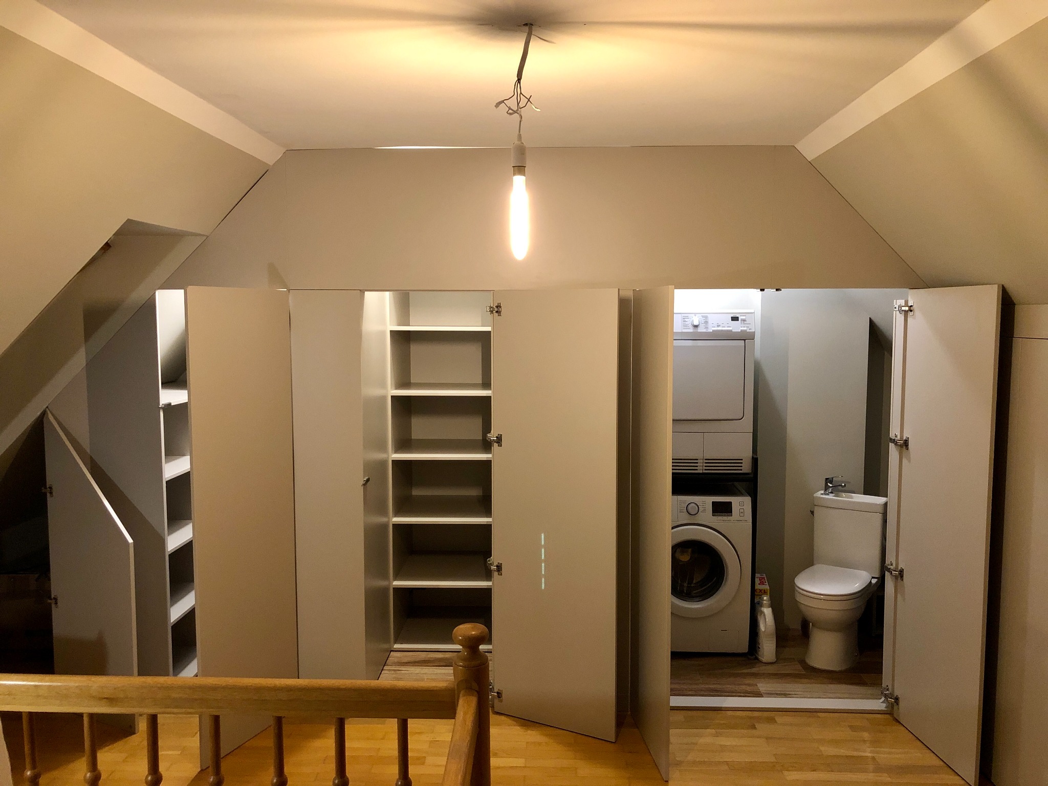 Garderoba na poddaszu z pralką i toaletą za drzwiami, oświetlona wiszącą żarówką, widok zza drewnianej balustrady.