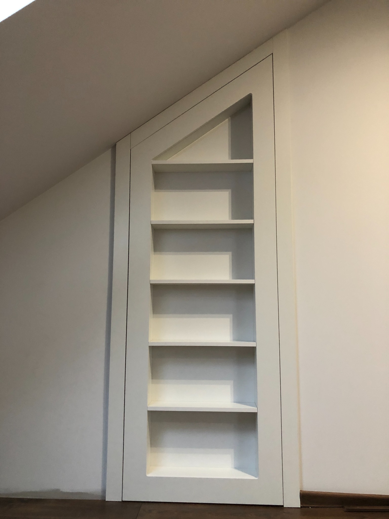 Biała, wbudowana półka z ukośną górą, dopasowana do skosu dachu, z pięcioma poziomami półek, na tle białej ściany i drewnianej podłogi.