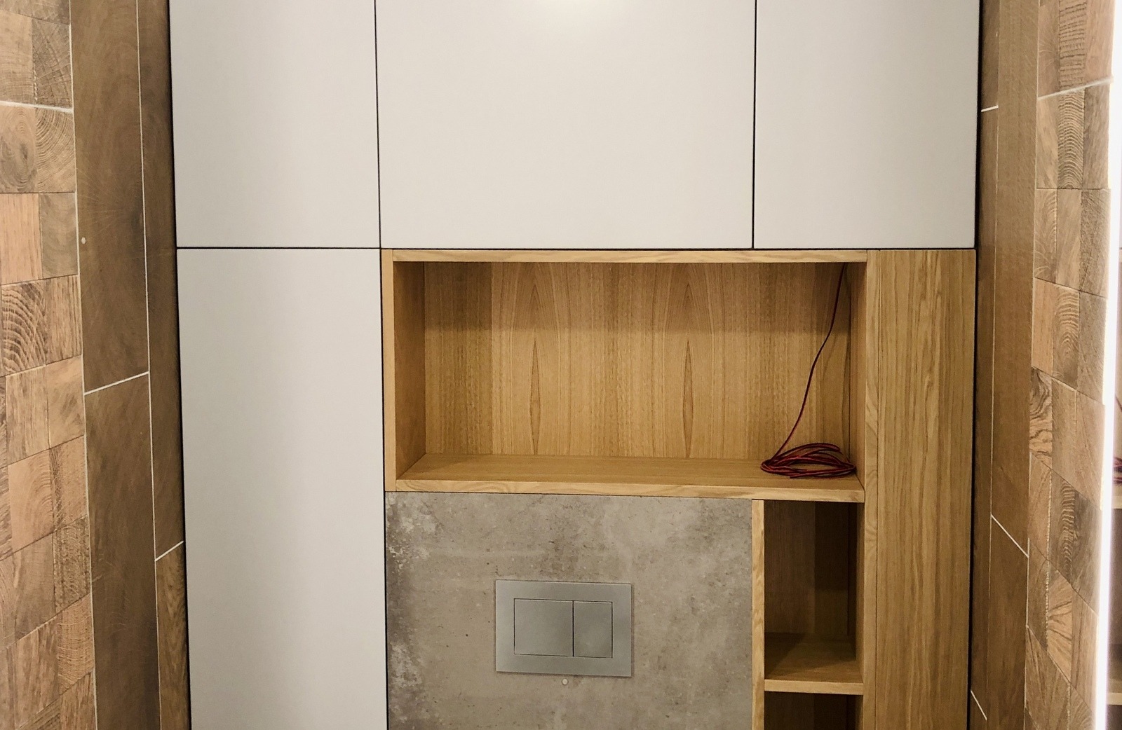 Białe i drewniane szafki łazienkowe z otwartą półką, nad zabudowaną spłuczką toaletową, na tle płytek imitujących drewno.