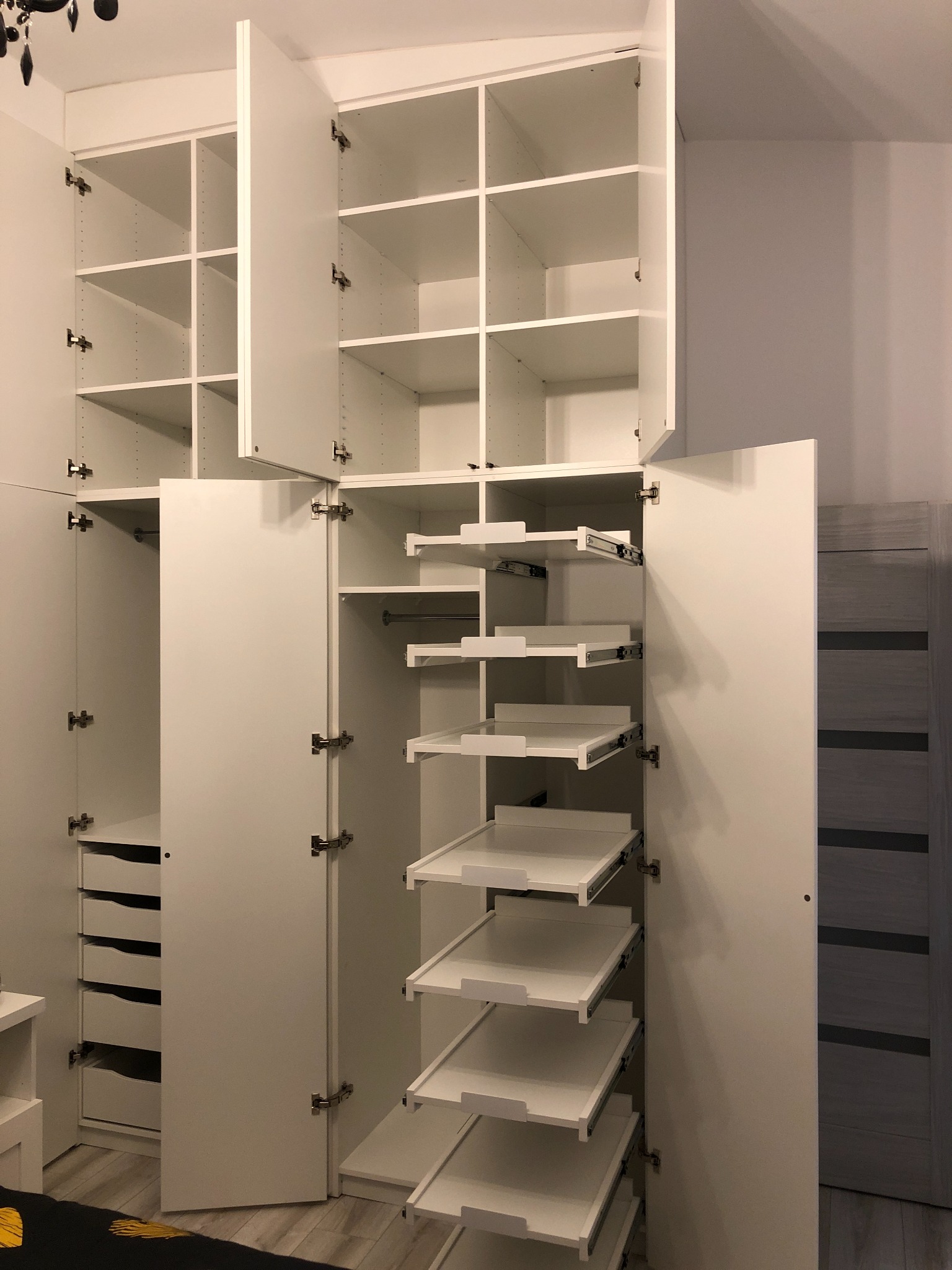 Wnętrze białej, otwartej garderoby z wysuwanymi półkami i szufladami, widoczne zawiasy i mocowania, oświetlenie dzienne.