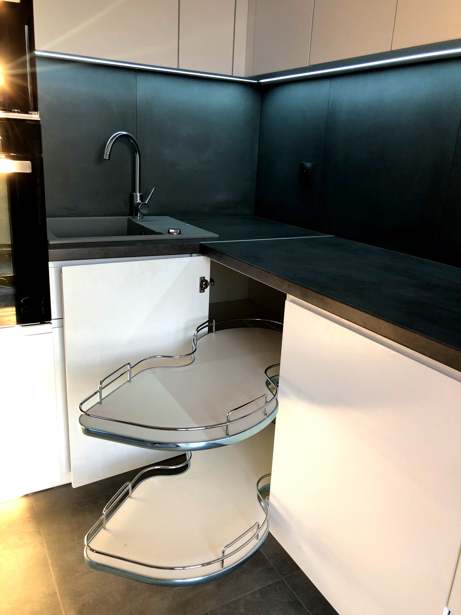 Narożna szafka kuchenna z otwartymi drzwiczkami, prezentująca wysuwane półki obrotowe z chromowanymi relingami, na tle czarnego blatu i ściany, z widoczną baterią zlewozmywakową.