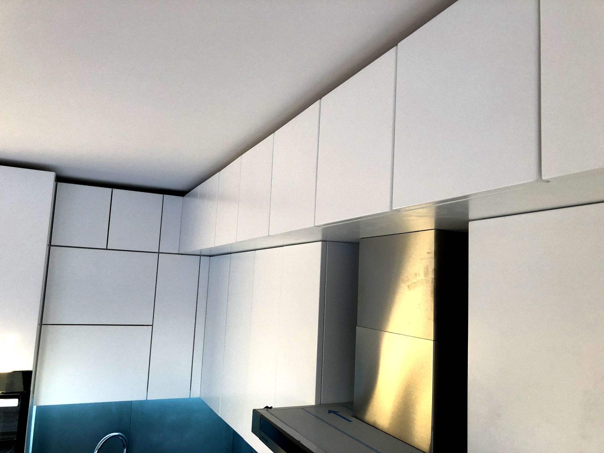 Białe, minimalistyczne szafki kuchenne pod sufitem, z widocznym okapem i fragmentem niebieskiego panelu nad zlewem.