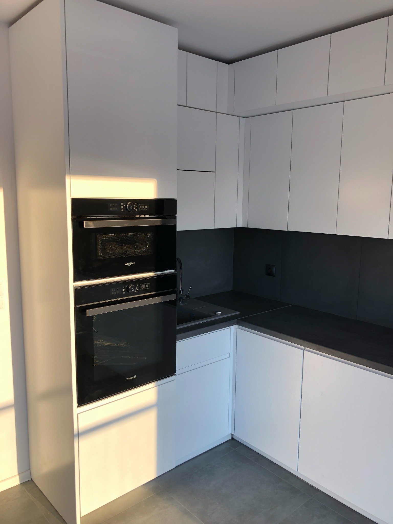 Nowoczesna kuchnia z białymi, gładkimi frontami, czarnym blatem i zabudowanymi piekarnikami Whirlpool, oświetlona popołudniowym słońcem.