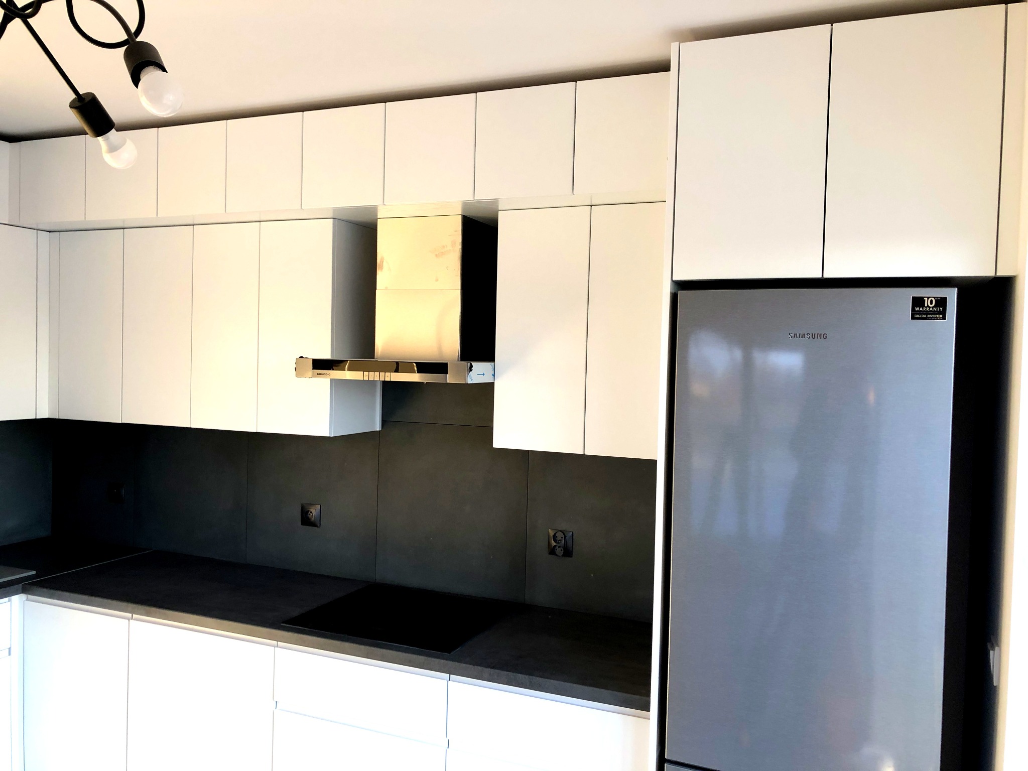 Nowoczesna kuchnia z białymi szafkami, czarnym blatem i szarym panelem ściennym, oświetlona lampą sufitową z odsłoniętymi żarówkami, obok stoi lodówka Samsung.
