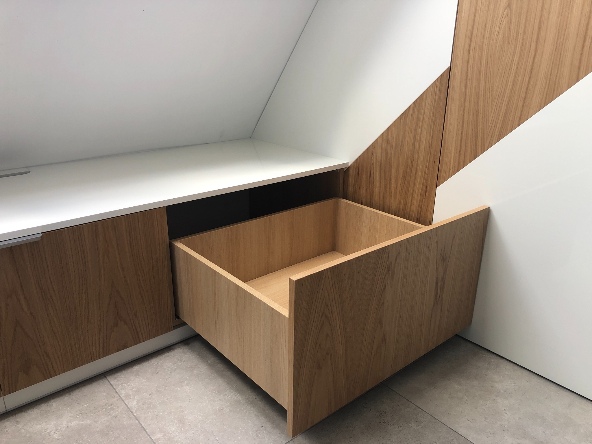 Otwarta szuflada z jasnego drewna w zabudowie pod skosem, biały blat i ściana, szare płytki podłogowe.