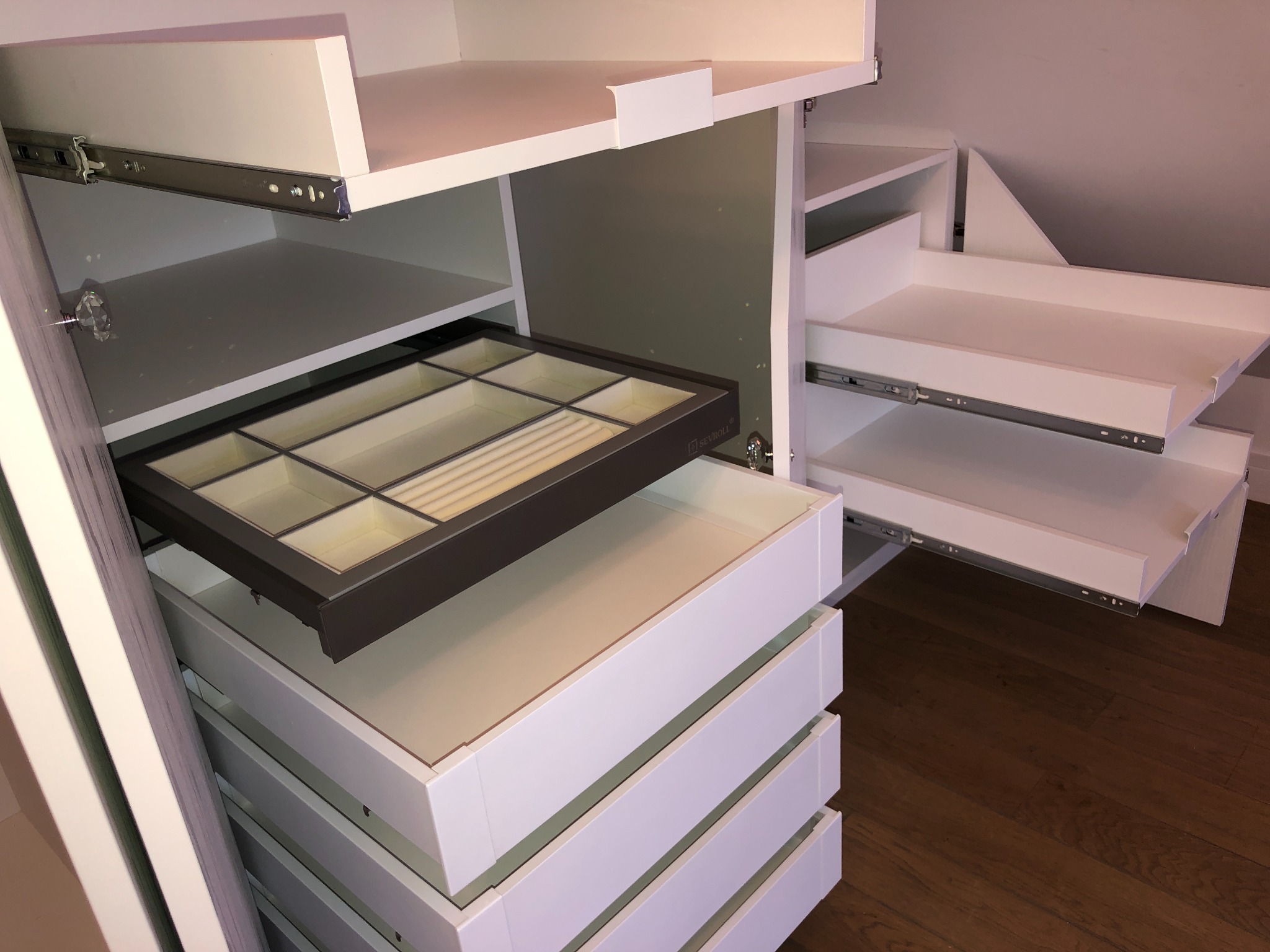 Biała szafa z wysuwanymi szufladami, z organizerem na biżuterię na górnej szufladzie, zamontowana na poddaszu ze skosami.