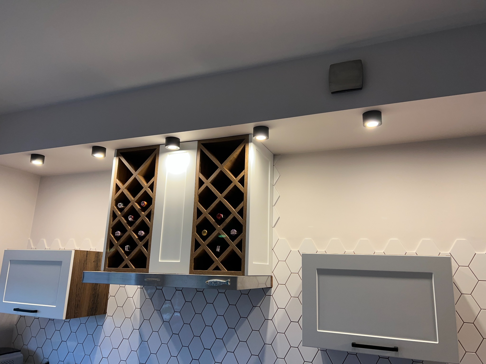 Nowoczesna zabudowa kuchenna z półkami na wino nad okapem, białe fronty szafek i heksagonalne płytki na ścianie.