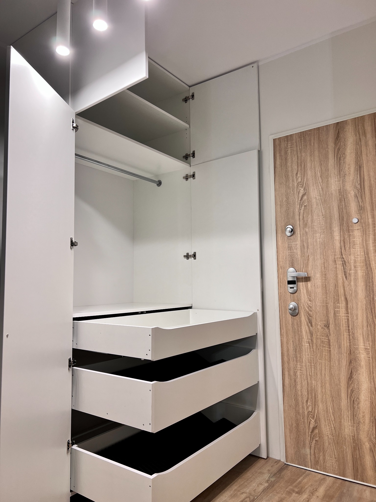 Biała szafa z otwartymi drzwiami i trzema wysuniętymi szufladami, obok drewnopodobnych drzwi wejściowych.