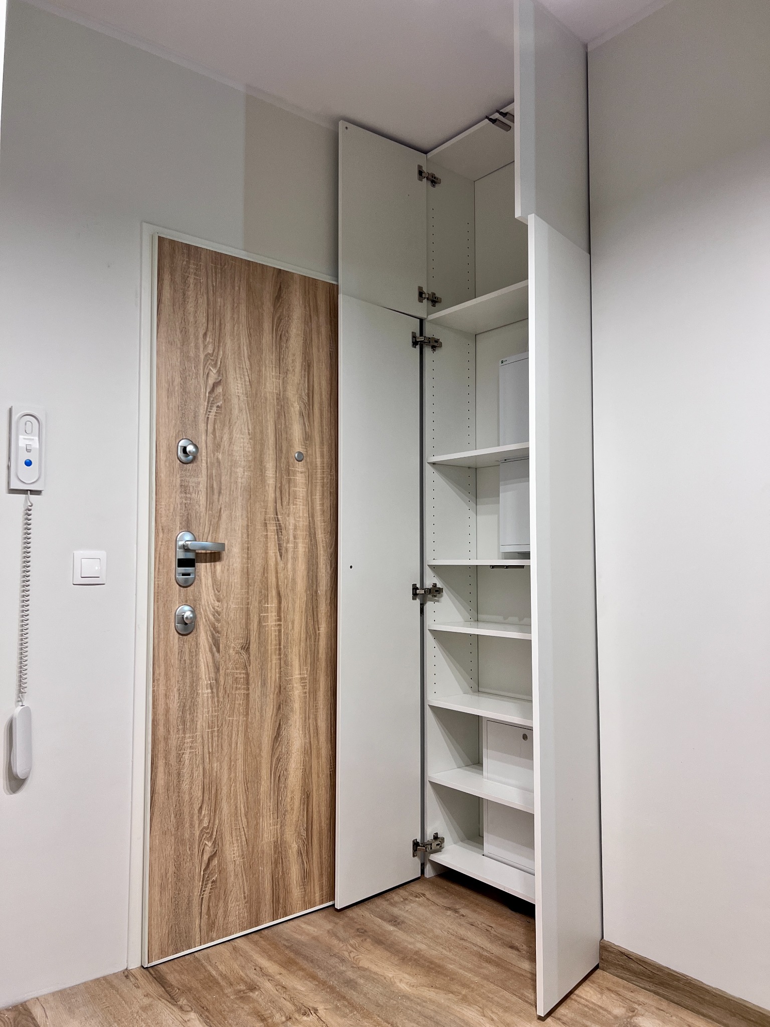 Biała szafa wnękowa z otwartymi drzwiami, widoczne półki, obok drzwi wejściowe z drewnopodobnym wykończeniem, minimalistyczne wnętrze w jasnych barwach.