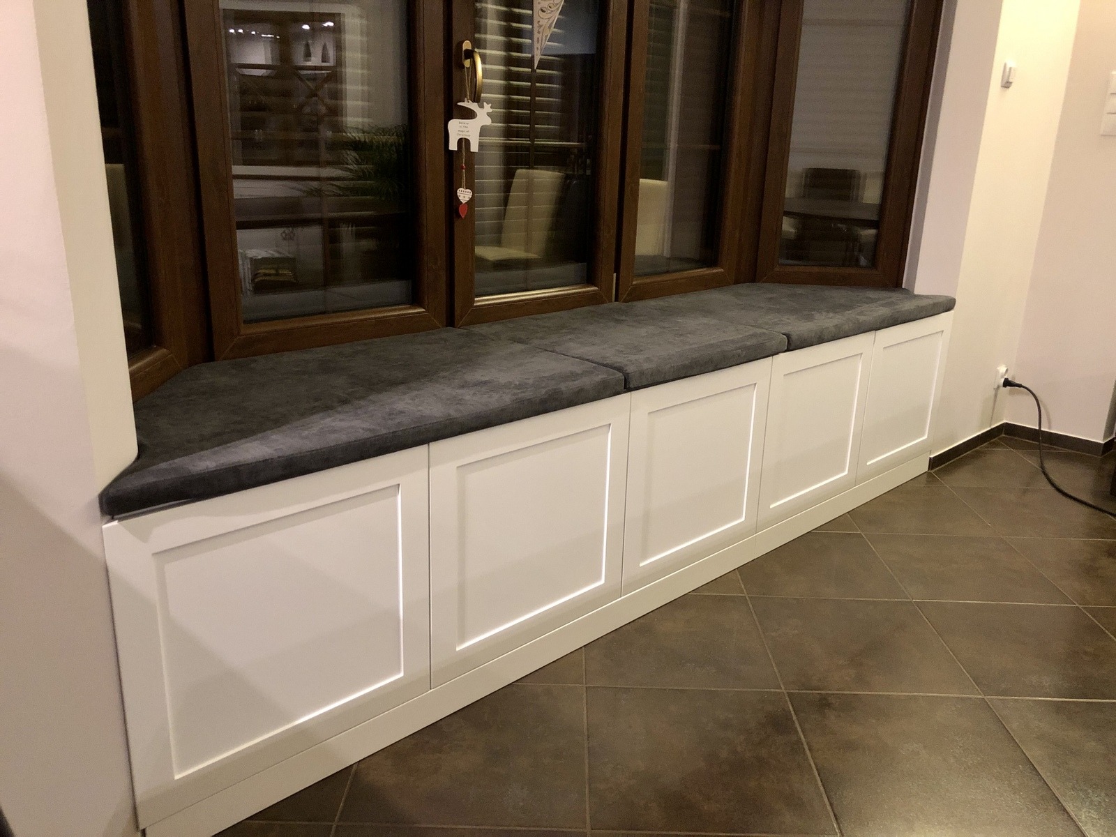 Zabudowa okna z siedziskiem z szarej tapicerki i białymi szafkami z frezowanymi frontami, widoczne brązowe ramy okienne i dekoracja świąteczna.