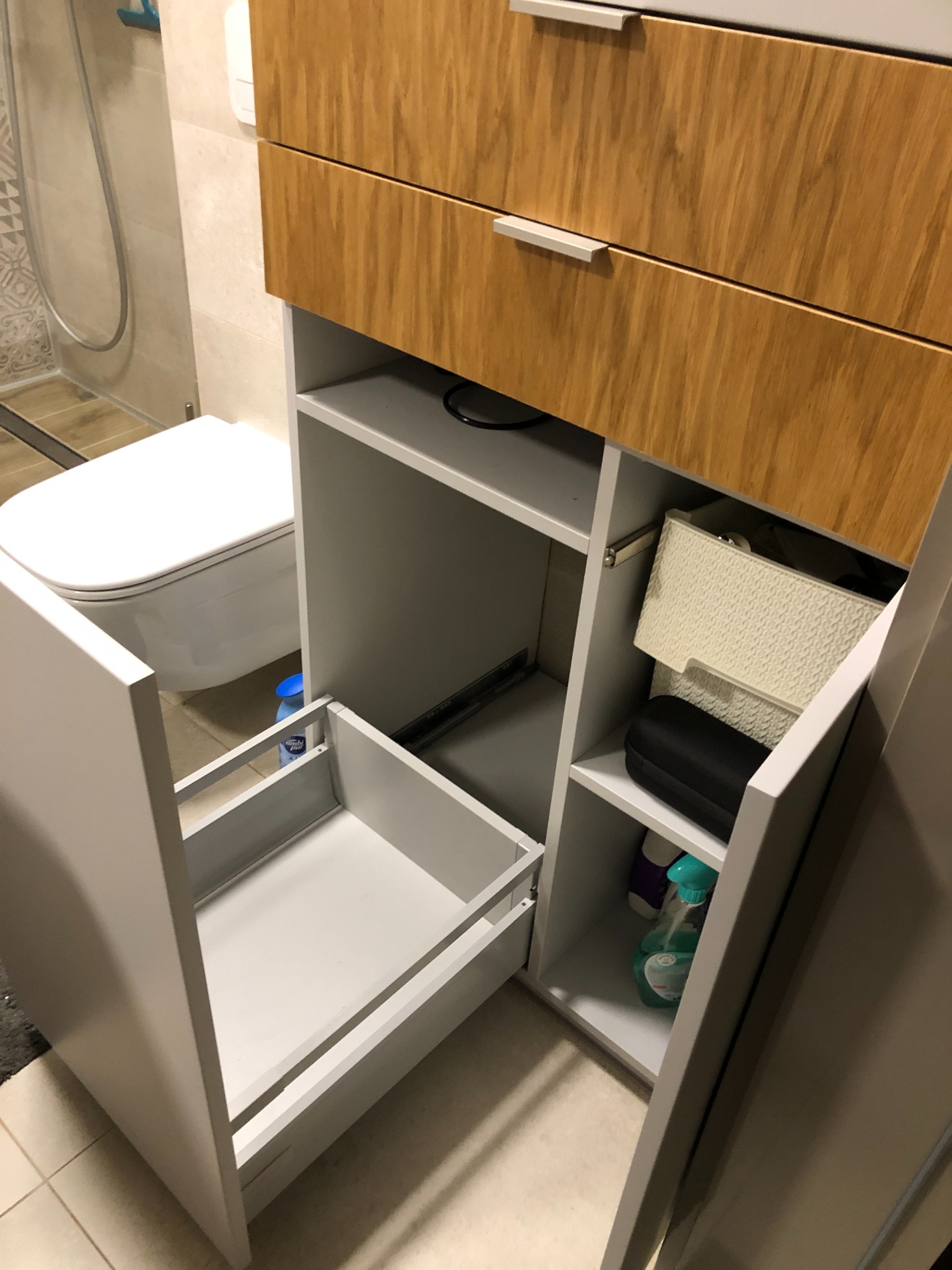 Szara szafka łazienkowa z drewnianymi szufladami i otwartymi półkami, widoczna otwarta szuflada i akcesoria łazienkowe na półkach, toaleta w tle.