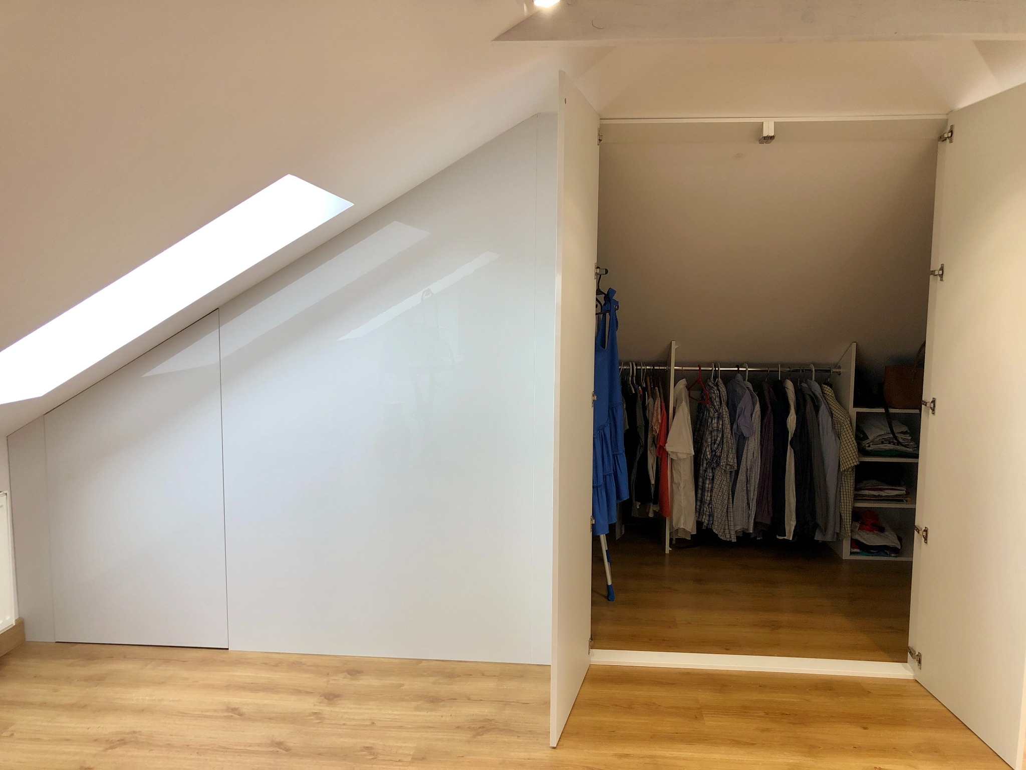 Biała zabudowa pod skosem z otwartą szafą wnękową, widoczne ubrania na wieszakach i półkach, drewnopodobna podłoga, okno dachowe.