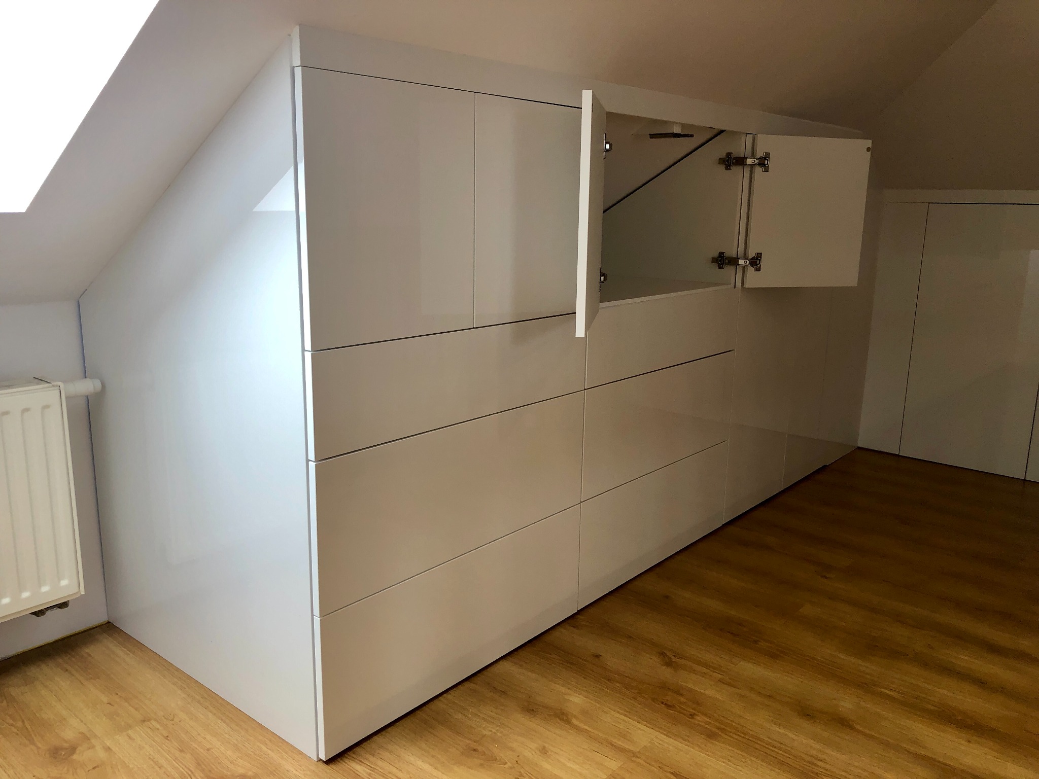 Biała, błyszcząca zabudowa meblowa ze szufladami i szafkami, dopasowana do skosu poddasza, z otwartymi drzwiczkami szafki ukazującymi zawiasy.