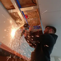 Pracownik w okularach ochronnych tnie metalową konstrukcję pod sufitem, iskry rozbryzgują się na boki, widoczne instalacje pod stropem.