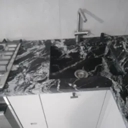 Czarny blat kuchenny z marmurkowym wzorem, z wbudowanym zlewem i płytą grzewczą; widoczny kran i białe szafki pod spodem.