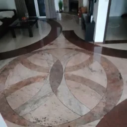 Wzorzysta podłoga z kamienia w salonie, z motywem dw&oacute;ch przeplatających się okręg&oacute;w w odcieniach beżu i brązu, widoczny kominek z kamienia i fragmenty mebli.