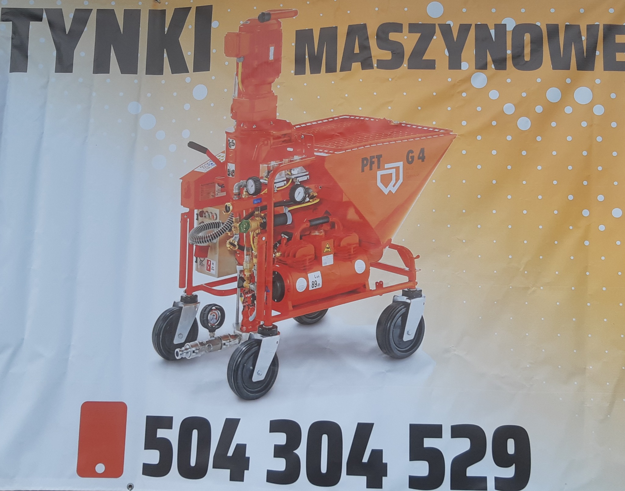 Pomarańczowa maszyna PFT G4 na kółkach, przeznaczona do nakładania tynków, widoczna na tle baneru reklamowego z numerem telefonu.