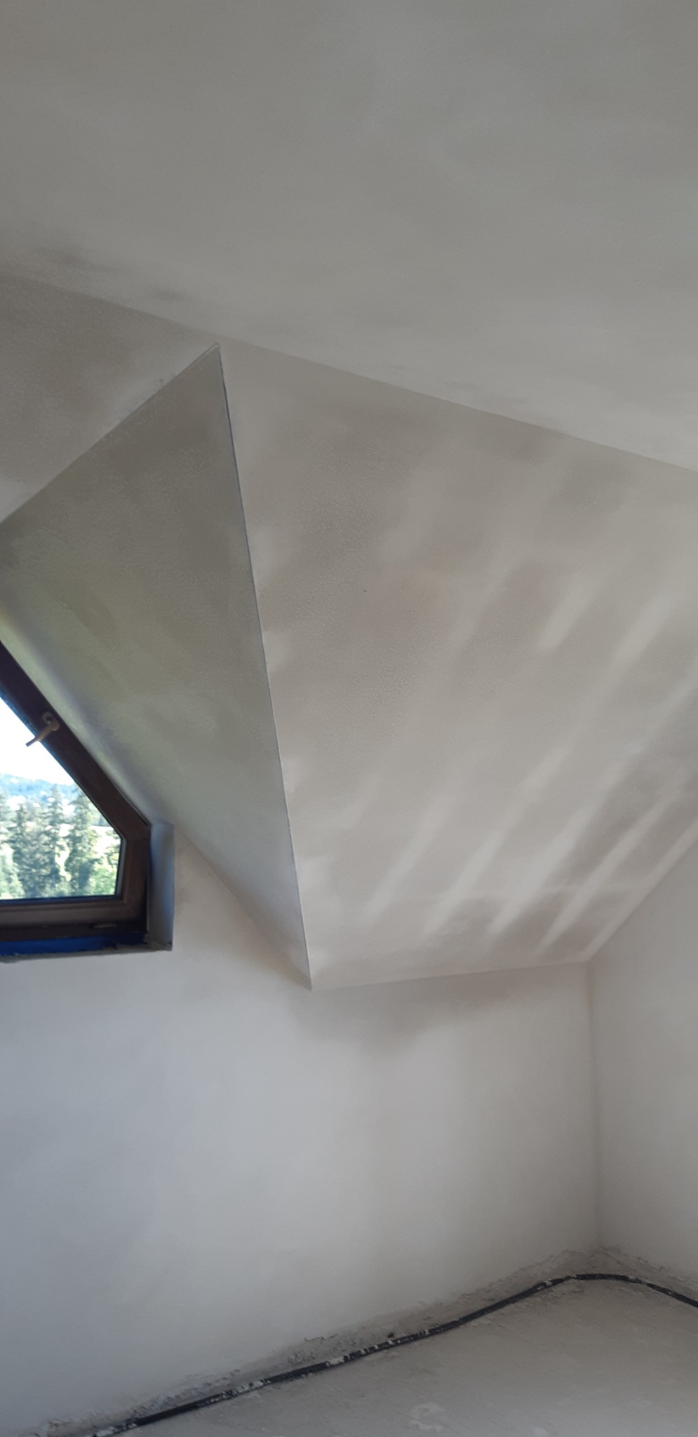 Poddasze po tynkowaniu maszynowym, widoczne okno z widokiem na zieleń, ślady po kablach przy podłodze.