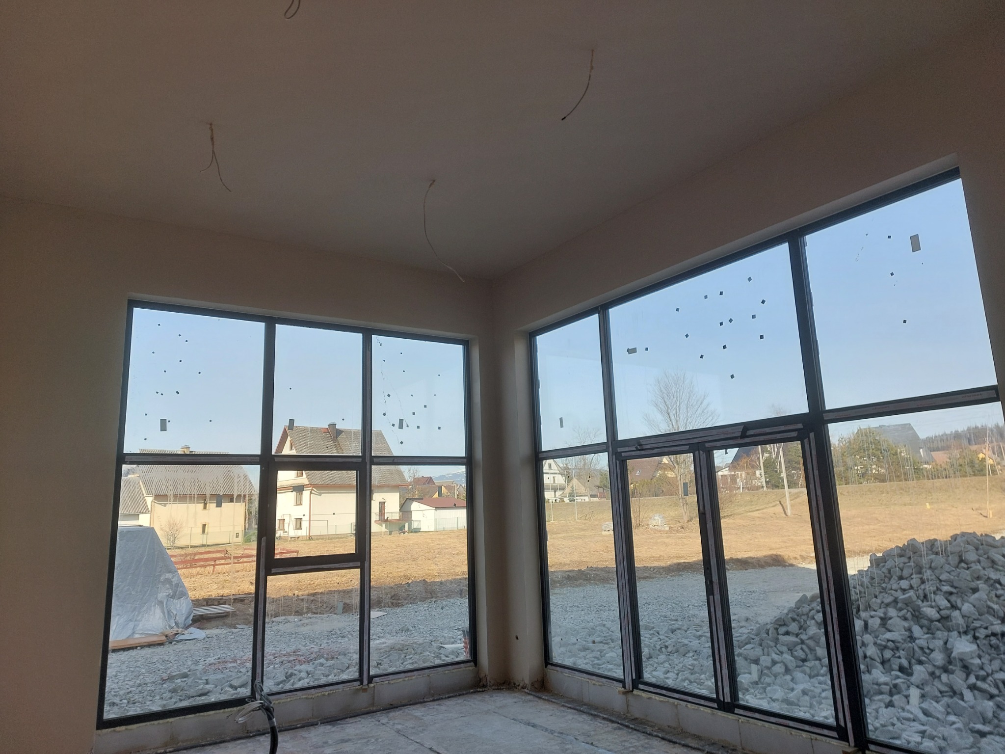 Wnętrze budynku w stanie deweloperskim z dużymi oknami, widoczne wyprowadzone kable oświetleniowe, ślady po naklejkach na szybach i przygotowane podłoże pod posadzkę.