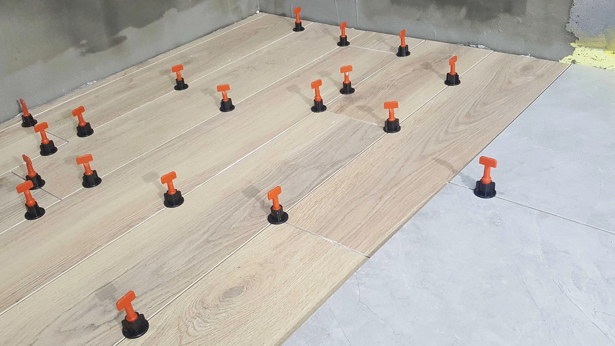 Układanie podłużnych płytek imitujących drewno z użyciem pomarańczowych klipsów poziomujących, widoczny fragment ściany z resztkami zaprawy i żółta pianka montażowa.