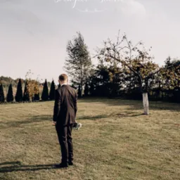 Mężczyzna w garniturze, stojący tyłem z bukietem na zielonej łące w jesiennej scenerii, z logo studia fotograficznego w górnej części kadru.
