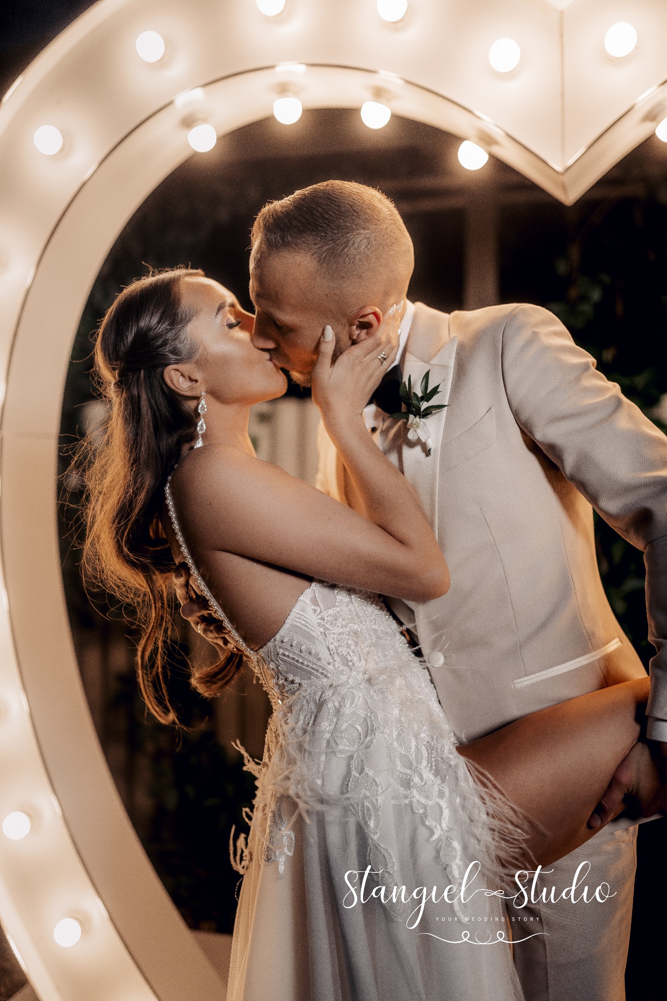 Pocałunek pary młodej na tle podświetlonego serca, panna młoda w sukni z koronkowymi zdobieniami i piórami, pan młody w beżowym garniturze z butonierką.