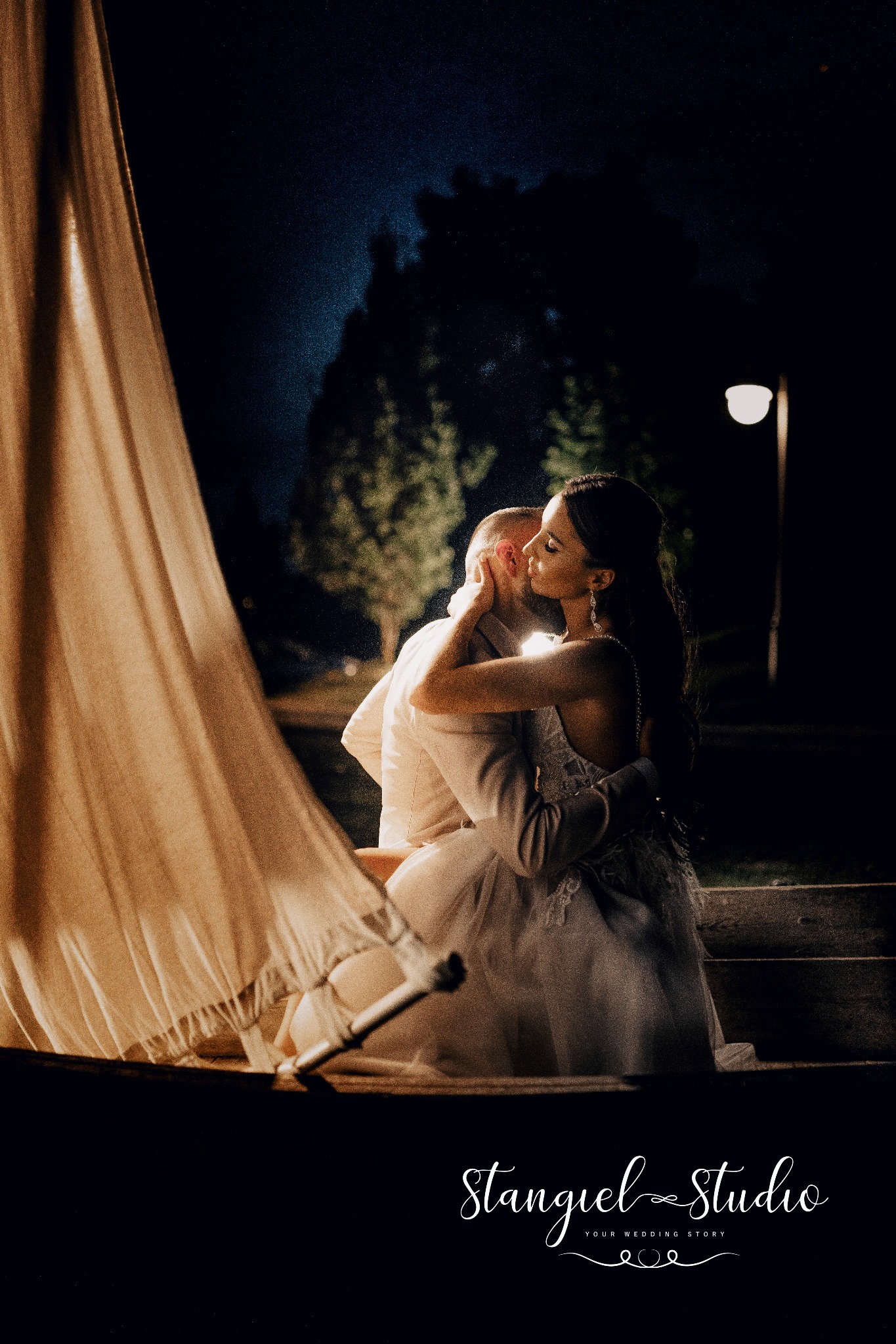 Para narzeczonych w objęciach w nocy, oświetlona lampą, z widocznym fragmentem żagla i logo studia fotograficznego w dolnej części kadru.