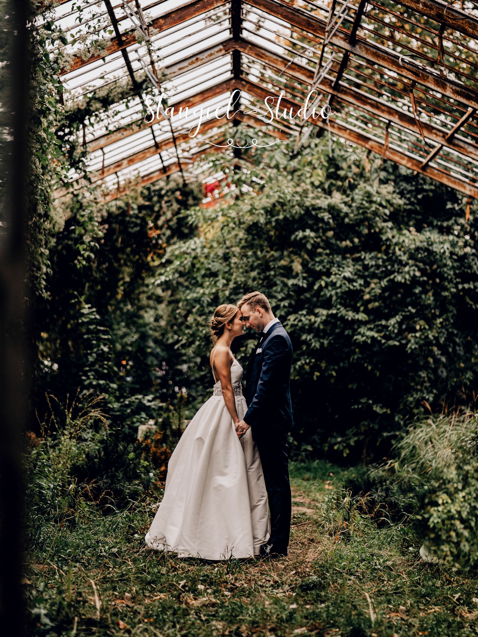 Para młoda trzymająca się za ręce w zarośniętej szklarni z rustykalną konstrukcją dachu.