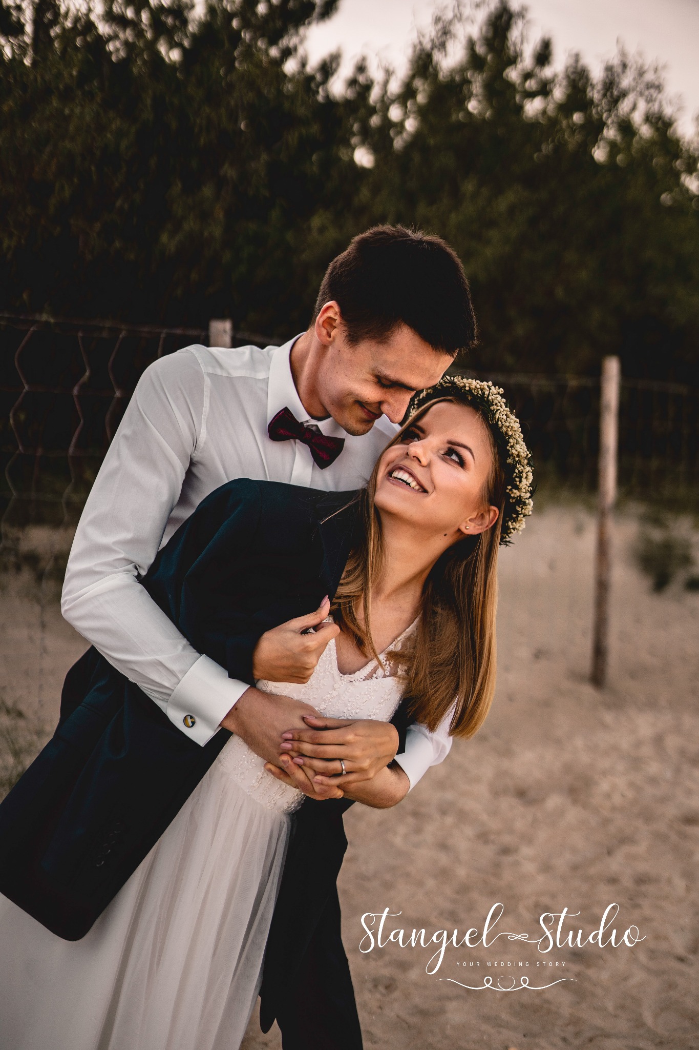 Uśmiechnięta para narzeczonych w objęciach na tle plaży i zarośli; ona w wianku z drobnych kwiatów, on w bordowej muszce i z marynarką przewieszoną przez ramię.