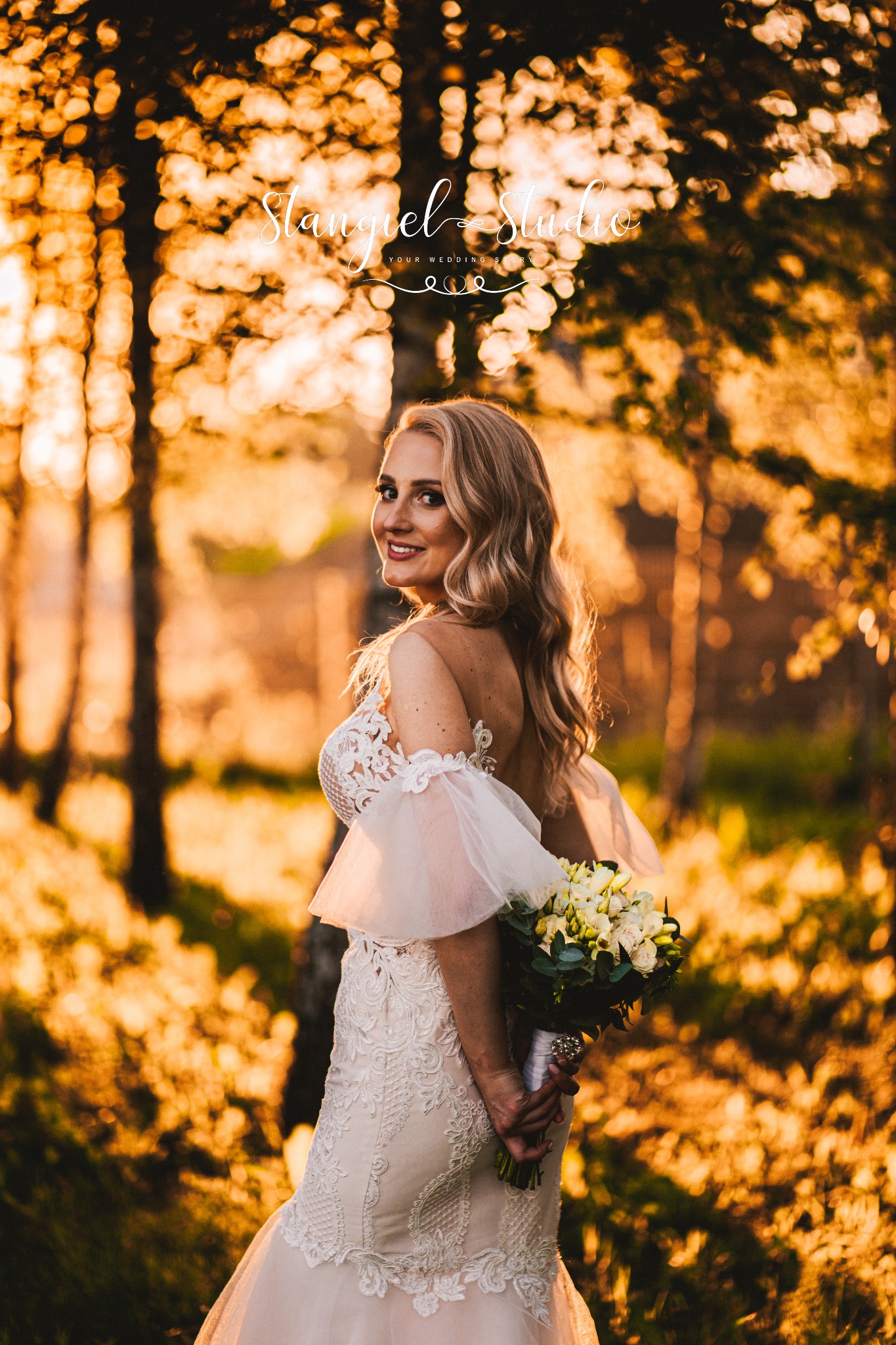 Uśmiechnięta panna młoda w koronkowej sukni, trzymająca bukiet, spogląda przez ramię w lesie oświetlonym ciepłym, zachodzącym słońcem. W tle widoczne logo studia.