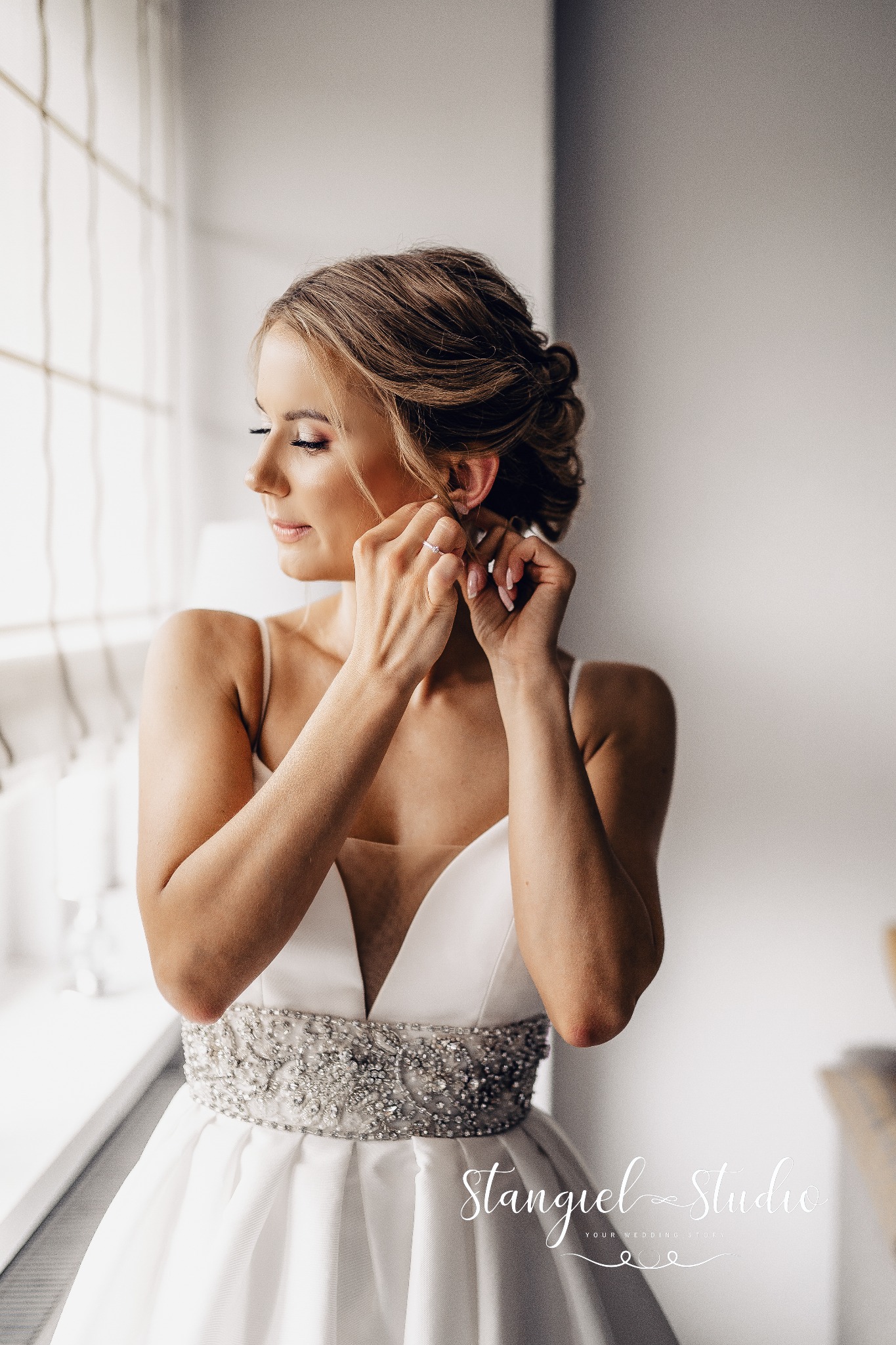 Panna młoda w białej sukni z kryształowym pasem, zakłada kolczyk przy oknie, profil, delikatny makijaż.
