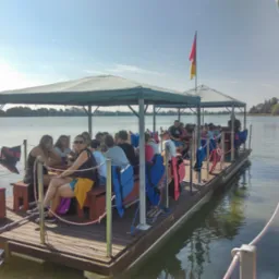 Grupa osób relaksuje się na pływającym pomoście z zadaszeniem, wyposażonym w ławki i silnik, na tle jeziora i drzew.