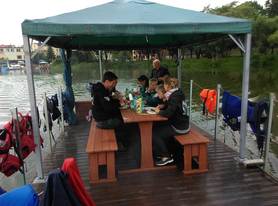 Rodzina je posiłek przy drewnianym stole na pływającej platformie z zadaszeniem na jeziorze, w tle budynki i drzewa, widoczne kamizelki ratunkowe.