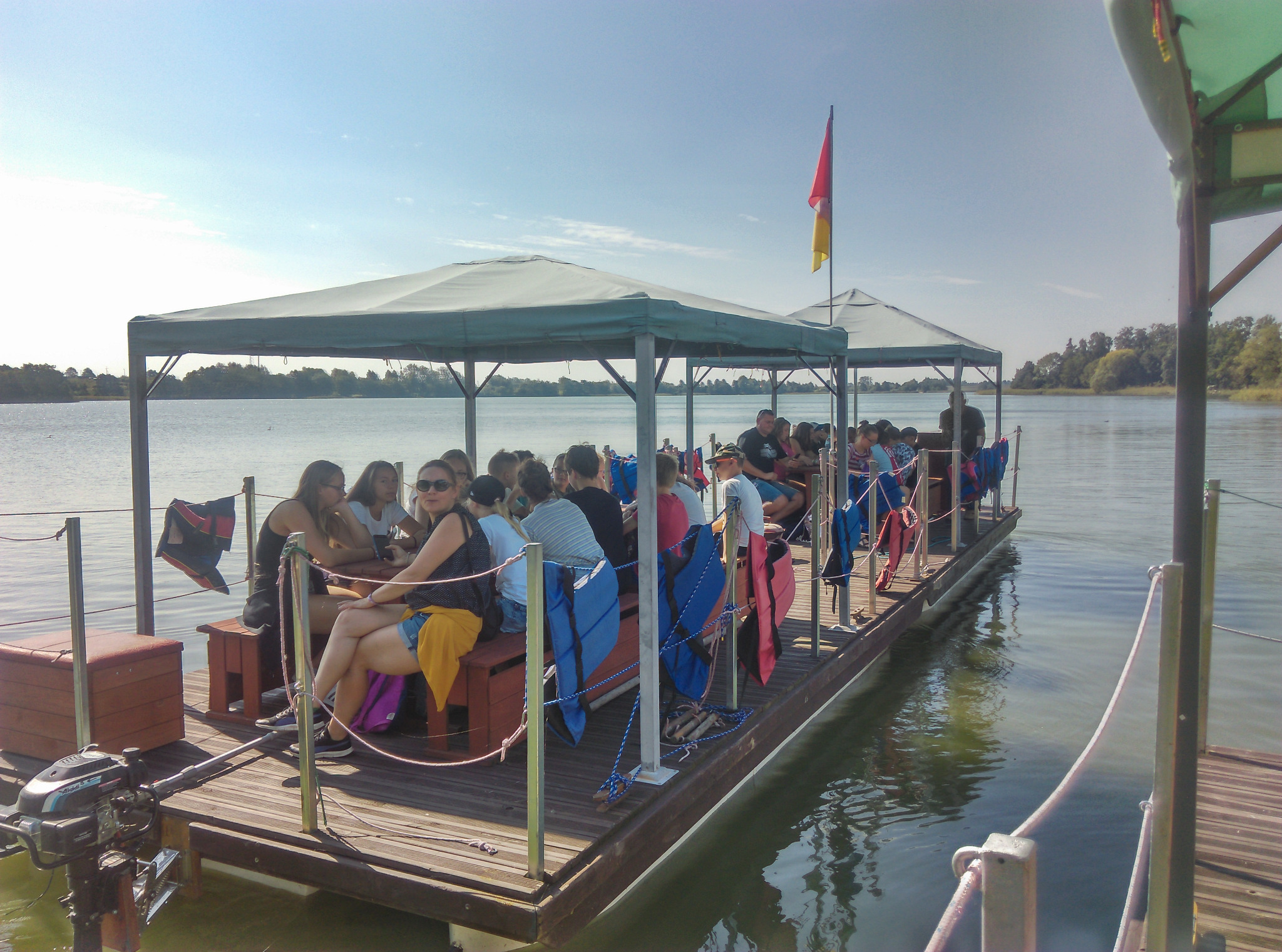 Grupa osób relaksuje się na pływającym pomoście z zadaszeniem, wyposażonym w ławki i silnik, na tle jeziora i drzew.