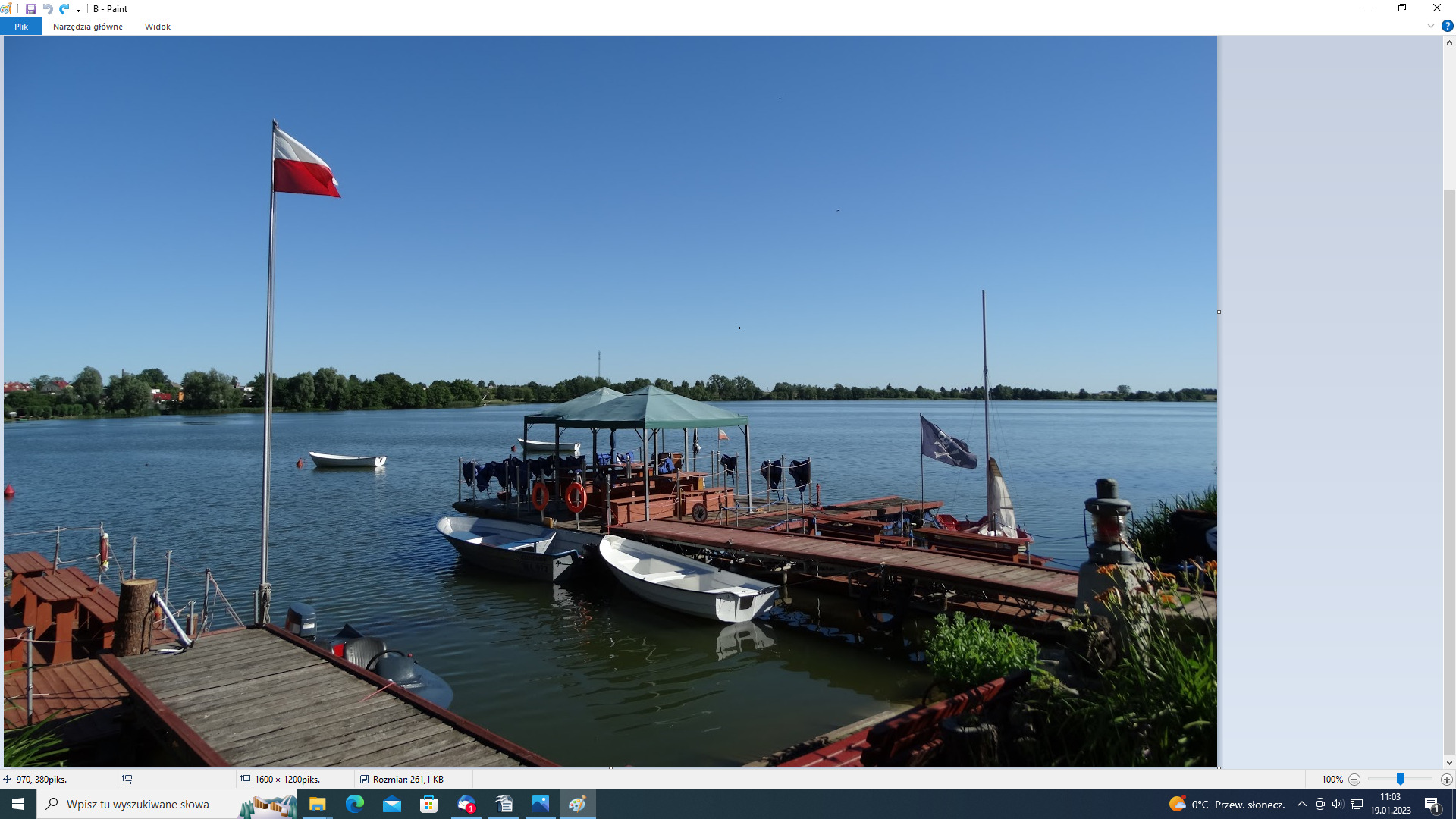 Przystań nad jeziorem z polską flagą, pomostem, łodziami i małą altaną z wyposażeniem ratunkowym.
