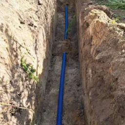 Wykopany rów z częściowo ułożoną niebieską rurą karbowaną, widok z góry w słoneczny dzień.