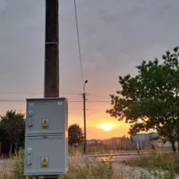 Szara skrzynka elektryczna z gniazdami na słupie betonowym na tle zachodzącego słońca, widoczne trawy i drzewa.