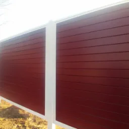 Długi, poziomy fragment ogrodzenia z desek w kolorze burgundowym, z białymi słupkami, widziany z niskiej perspektywy w słoneczny dzień.