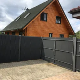 Nowoczesna, antracytowa brama wjazdowa z metalową furtką i skrzynką pocztową na tle drewnianego domu jednorodzinnego w Moryniu, widok z kostki brukowej.
